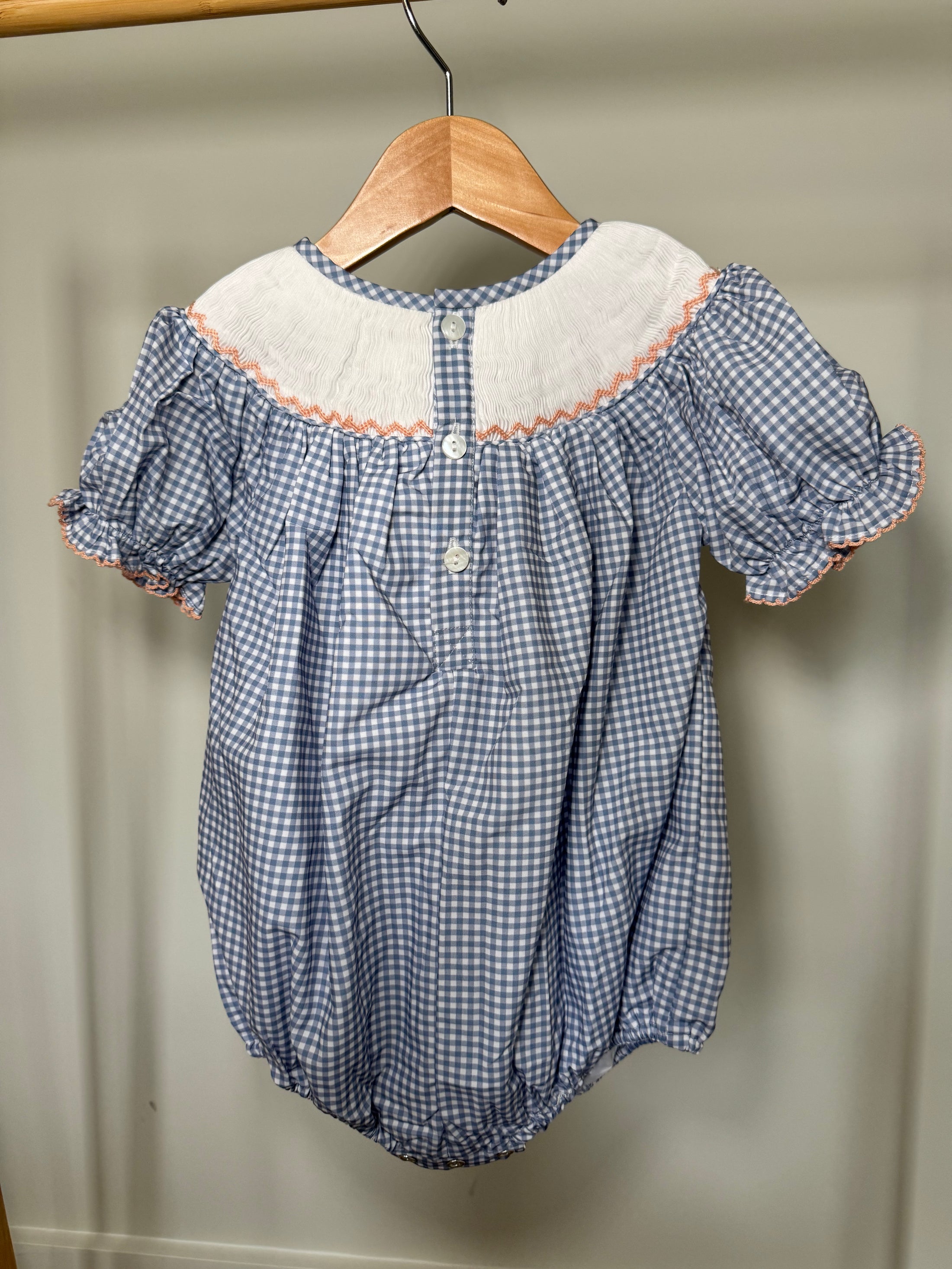 Blue checkered baby romper with white collar on a wooden hanger against a beige wall.
